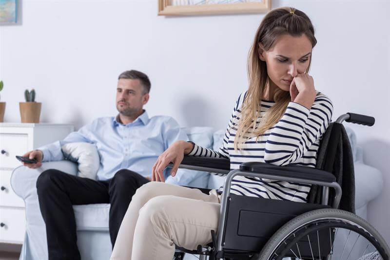 Sad injured woman sitting on wheelchair while her husband is on the sofa.