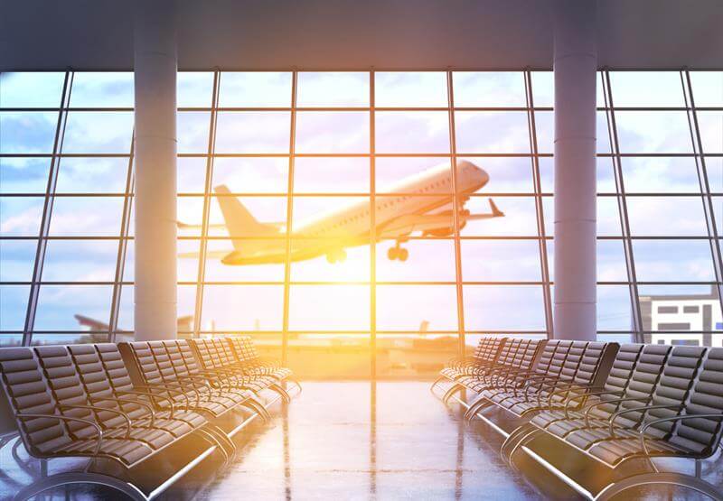Airport terminal interior with rows of seats, framed windows with city view, sunlight and an airplane flying.